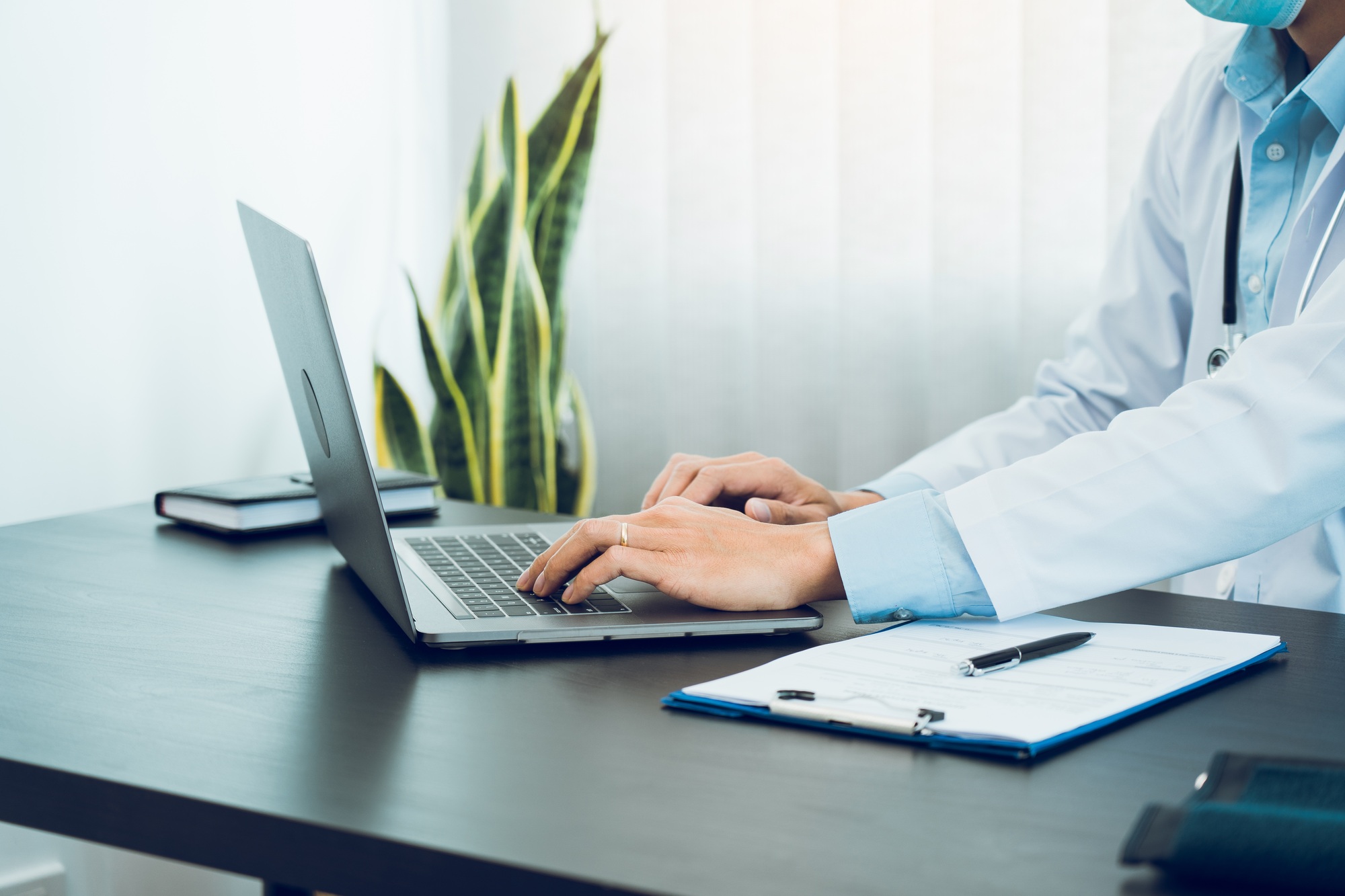 Doctor was sitting in the office working on a laptop computer while wearing a mask.